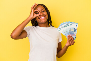 Young african American woman holding bills isolated on yellow background  excited keeping ok gesture on eye.