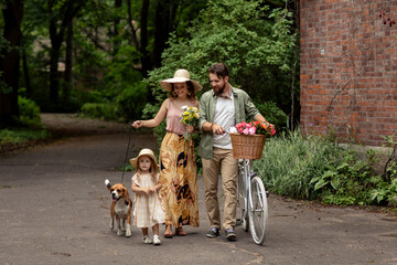 Beautiful caucasian family walking in park with vintage bicycle with basket of flowers, daughter and beagle. 