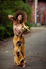 Full length portrait of beautiful smiling young romantic caucasian woman with bouquet of flowers in straw hat at sunset in summer outdoor. Sun rays in hair.