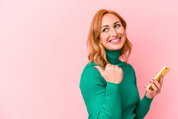 Young caucasian woman holding a mobile phone isolated on pink background points with thumb finger away, laughing and carefree.