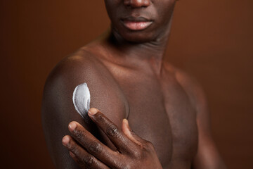 Close-up of African young man applying cream on his body for protection isolated on brown background