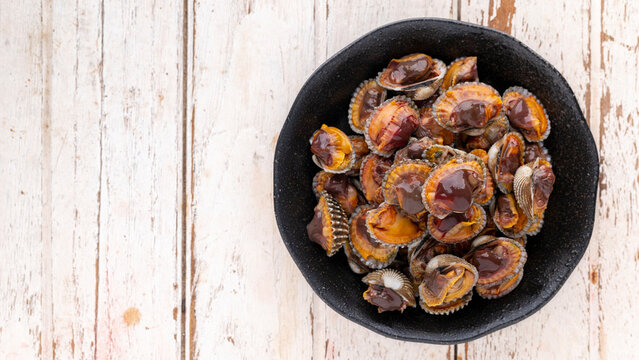 fresh steamed cockles, boiled cockles in black ceramic plate on white wood texture background with copy space for text, top table view, flat lay, blood cockle