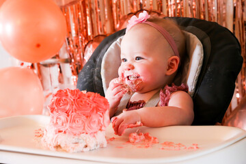 Cute birthday girl celebrating her one year birthday by messily eating a cake surrounded by pink background streamers and balloons