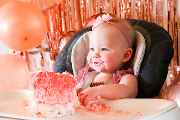 Cute birthday girl celebrating her one year birthday by messily eating a cake surrounded by pink background streamers and balloons