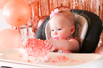 Cute birthday girl celebrating her one year birthday by messily eating a cake surrounded by pink background streamers and balloons