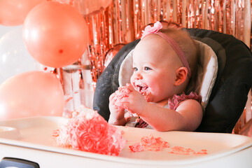 Cute birthday girl celebrating her one year birthday by messily eating a cake surrounded by pink background streamers and balloons