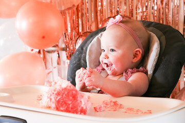 Cute birthday girl celebrating her one year birthday by messily eating a cake surrounded by pink background streamers and balloons