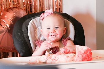Cute birthday girl celebrating her one year birthday by messily eating a cake surrounded by pink background streamers and balloons
