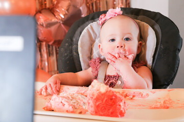Cute birthday girl celebrating her one year birthday by messily eating a cake surrounded by pink background streamers and balloons