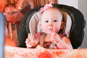 Cute birthday girl celebrating her one year birthday by messily eating a cake surrounded by pink background streamers and balloons