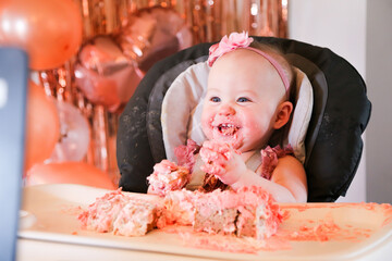 Cute birthday girl celebrating her one year birthday by messily eating a cake surrounded by pink background streamers and balloons