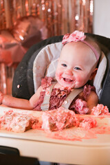 Cute birthday girl celebrating her one year birthday by messily eating a cake surrounded by pink background streamers and balloons