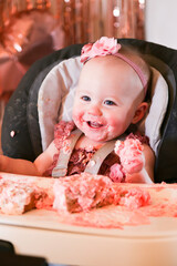 Cute birthday girl celebrating her one year birthday by messily eating a cake surrounded by pink background streamers and balloons