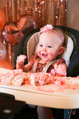 Cute birthday girl celebrating her one year birthday by messily eating a cake surrounded by pink background streamers and balloons