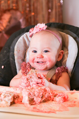 Cute birthday girl celebrating her one year birthday by messily eating a cake surrounded by pink background streamers and balloons
