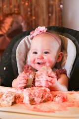 Cute birthday girl celebrating her one year birthday by messily eating a cake surrounded by pink background streamers and balloons