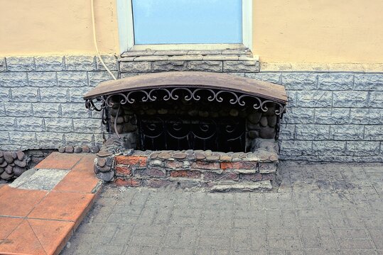 old window in the basement with an iron grate on a brick brown wall near the gray pavement on the street