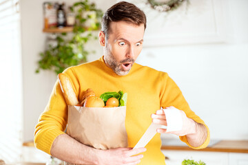 Surprised man looking at store receipt after shopping, holding a paper bag with healthy food. Guy...