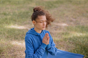 a European woman with curly hair meditates in a park. yoga and meditation for mental and physical health. a woman sits in the lotus position and namaste with her eyes closed and engages in breathing