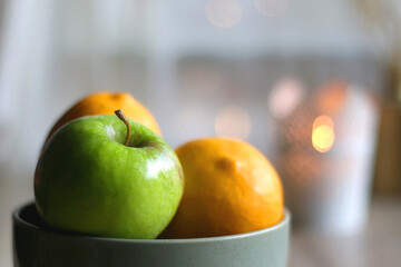 Bowl of lemons and apples, candle holder with lit candle and vase with gypsophila flowers on the table. Bokeh lights in the background. Selective focus.