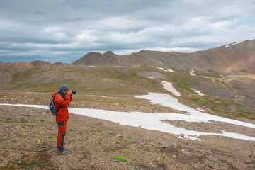 Obraz premium Atmospheric scenery with man in red against mountain range with sharp rocks under gray cloudy sky. Tourist with camera shoots dramatic mountain landscape. Man photographs mountains at gloomy weather.
