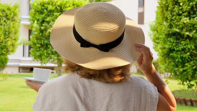Back view of a woman in a straw hat with a white cup of hot drink of coffee in her own garden on the terrace in summer with a sunny day. Morning concept, morning enjoyment, tune in for a happy day
