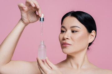 Asian woman with naked shoulders holding dropper and serum isolated on pink