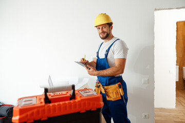Smiling handyman worker, builder in uniform and hard hat looking at camera, making notes on a clipboard while standing indoors