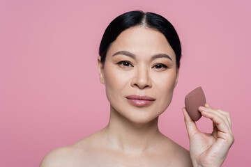 Asian woman with naked shoulders holding beauty blender isolated on pink