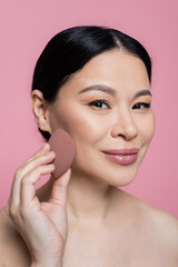 Smiling asian woman applying face foundation with beauty blender isolated on pink