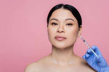 Cosmetologist in latex glove holding syringe near face of asian woman isolated on pink