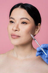 Cosmetologist holding syringe near face of asian woman isolated on pink