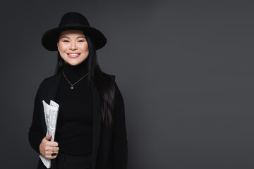 Smiling asian woman in jacket and fedora hat holding newspaper isolated on dark grey