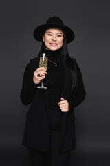 Fototapeta na wymiar Positive asian woman in fedora hat holding champagne isolated on dark grey