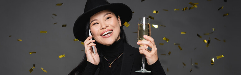 Happy asian woman in fedora hat talking on smartphone and holding champagne under confetti on dark...
