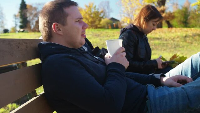 a man and a woman are sitting on a park bench. a woman is flipping through a tape on her phone, a man is drinking tea from a mug. autumn family vacation