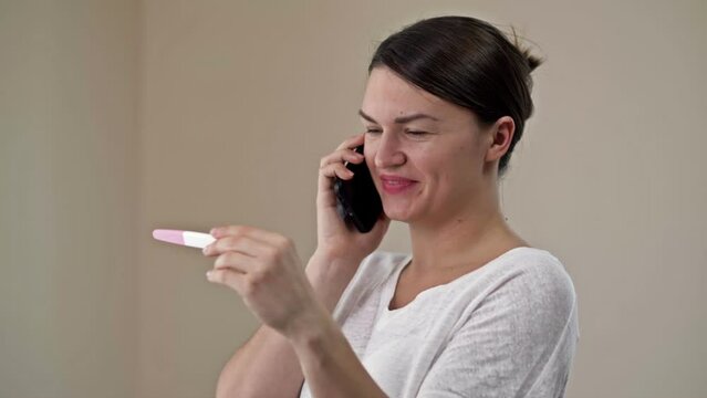 Happy woman holding her positive result on pregnancy test and talking by a smartphone with the family.