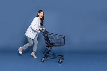 Full length portrait of young Asian woman pushing an empty shopping cart or shopping trolley isolated on deep blue background - 485621153