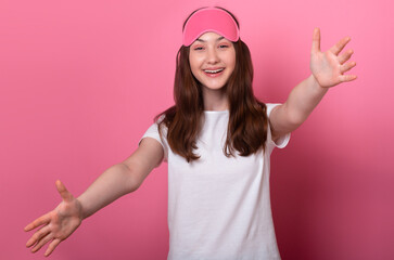 Obraz na płótnie Canvas happy teen girl gives hug at camera spreads hands calling a pet, want to embrace parents mom and dad to wish good night wearing a sleep mask and a white t-shirt on pink background home online learning