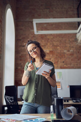 Smiling woman, interior designer or architect in casual wear with messy hairdo holding pen and notebook, pointing at camera, working on new project, standing in her office