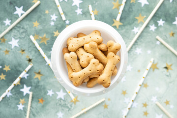 Dog treats, bone biscuits in white bowl on festive green background with gold and white stars and cocktail straws. Pet birthday party. Top view, selective focus. Care and love for puppy. Dog Day.