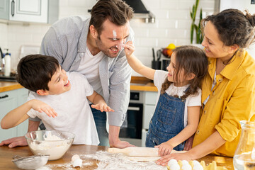 Fototapeta premium Happy family of four cooking in the kitchen together, parents and two kids making dough together, preparing pie or cookie, have a fun on weekend, daughter smearing nose of dad with flour and laughing