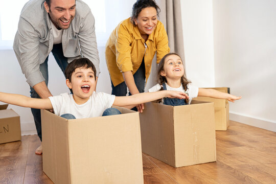 Multiracial family of four have fun in new house, parents riding kids in cardboard boxes in empty living room, happy children spreading arms and laughing. Family moving in new home, relocation concept