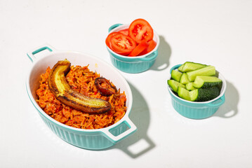 Jollof rice with fried banana. Fresh vegetables - tomato and cucumber. White background. Traditional Nigerian rice food with tomatoes, onions and spices.