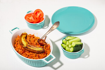 Jollof rice with fried banana. Fresh vegetables - tomato and cucumber. White background. Traditional Nigerian rice food with tomatoes, onions and spices. Empty plate in turquoise color.