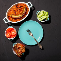 Jollof rice with fried banana, baked chicken. Rice with tomatoes, onions, spices. Traditional national Nigerian food. Dark background. View from above. Empty plate in turquoise color.