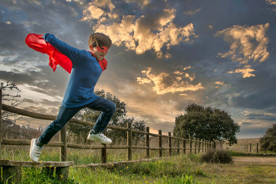 Child Superhero Portrait, Power Concept.
Little Boy Who Pretends To Be A Superhero, Wears A Red Mask And A Red Cape And Is In The Middle Of Nature. Image With Copy Space