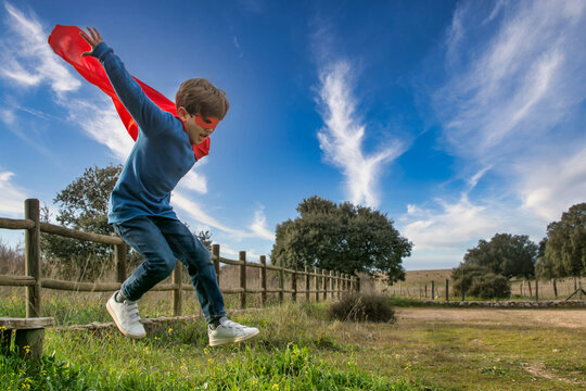 Child Superhero Portrait, Power Concept.
Little Boy Who Pretends To Be A Superhero, Wears A Red Mask And A Red Cape And Is In The Middle Of Nature. Image With Copy Space