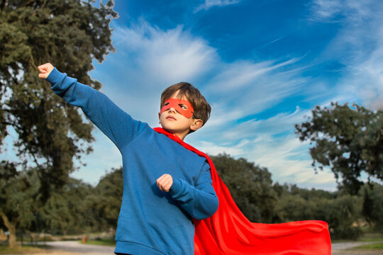 Child Superhero Portrait, Power Concept.
Little Boy Who Pretends To Be A Superhero, Wears A Red Mask And A Red Cape And Is In The Middle Of Nature. Image With Copy Space