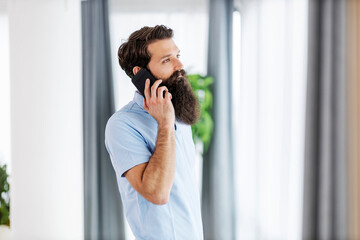 Serious bearded man standing at his home and having phone call.